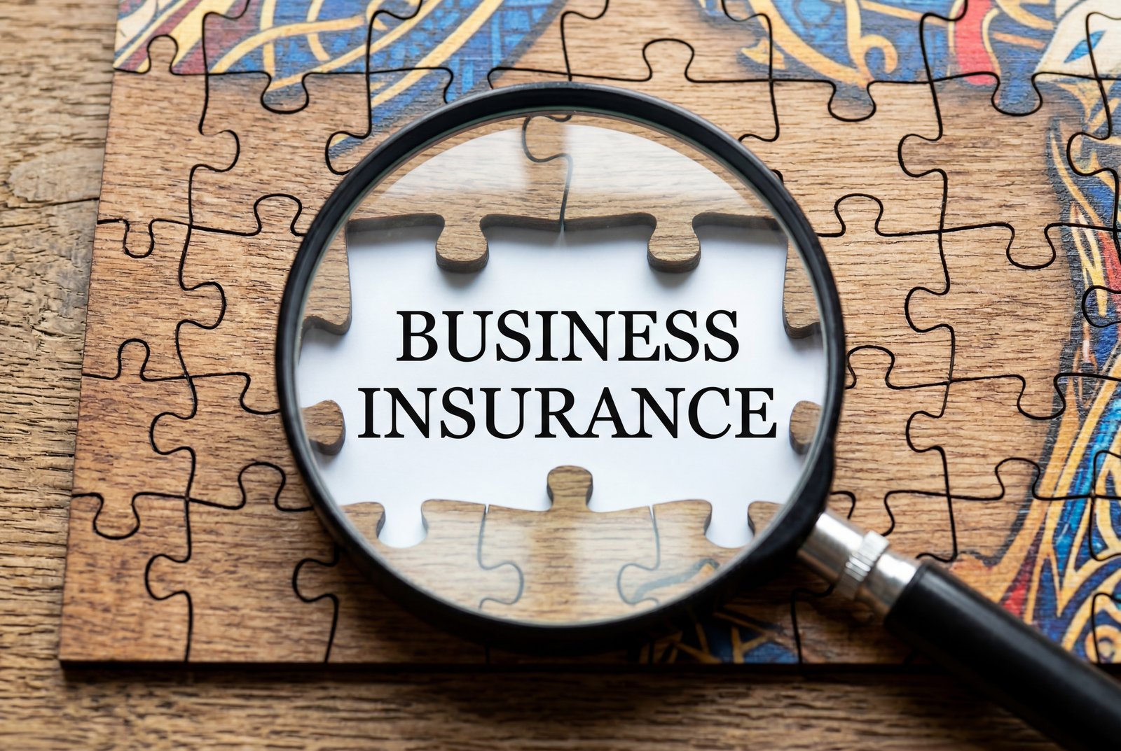 A magnifying glass focusing on the words BUSINESS INSURANCE printed on a white puzzle piece missing from a wooden jigsaw puzzle