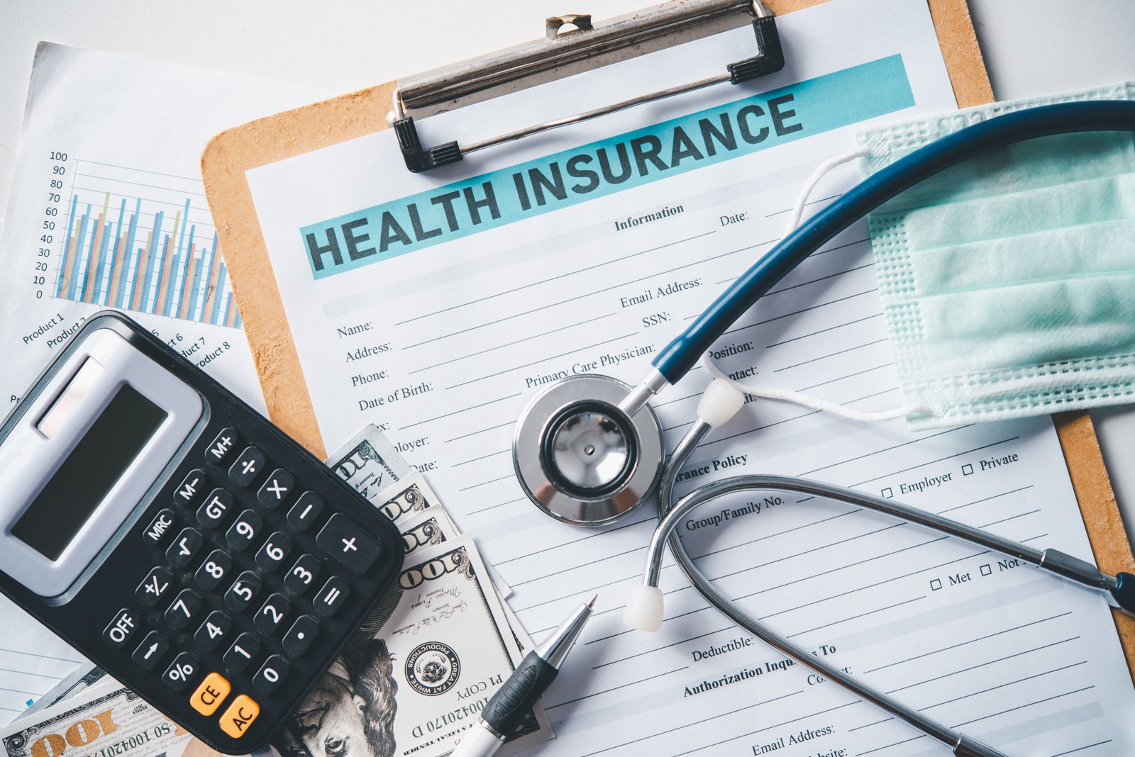 A printed Health Insurance application form clipped to a clipboard, surrounded by a stethoscope, a calculator, a surgical mask, a pen, and paper cash